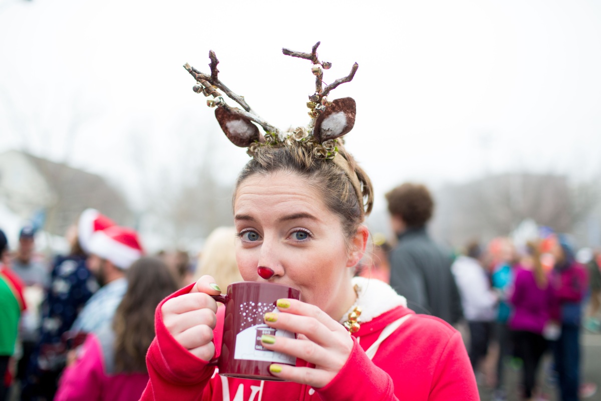 A woman wearing antlers and a red nose sips hot chocolate from a red mug. A crowd wears red hats in the background.