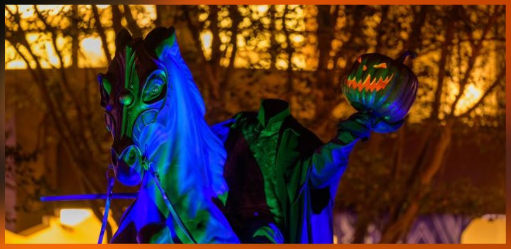 A person in a dark jacket holds a pumpkin on a stick in front of a brightly lit background during nighttime festivities.