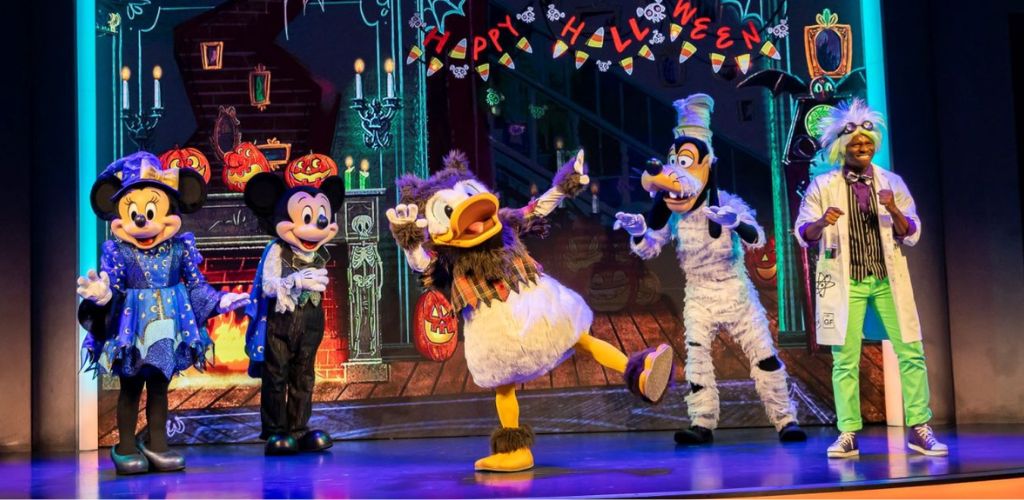 Costumed characters including Mickey Mouse Minnie Mouse Donald Duck Goofy and a performer in a white coat pose on stage with Halloween decorations and a festive background.
