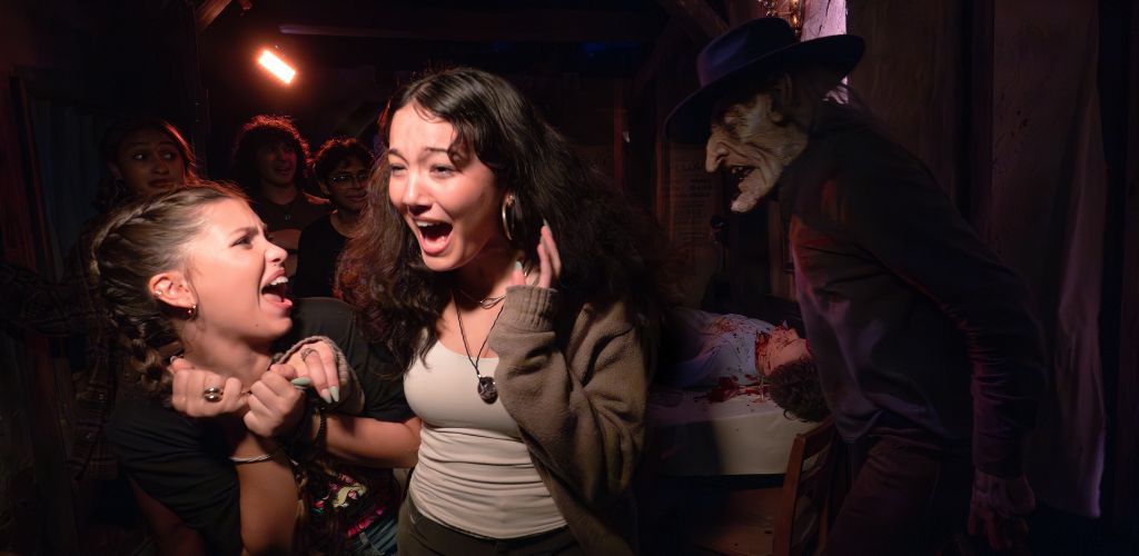 People in costumes scare and surprise each other at a Halloween party with spooky decorations and a fake bloody scene.