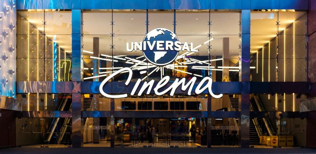 Universal Cinema theater entrance with illuminated signage and modern architectural design, featuring glass doors and escalators inside.