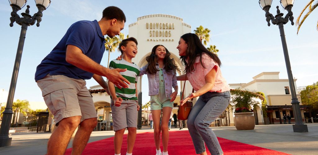 A family of four smiling and dancing on a red carpet in front of Universal Studios Hollywood during daytime.