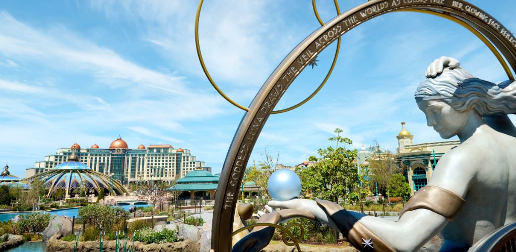 A large statue of a woman with flowing hair holding a spherical object in her hand, set in a colorful landscaped outdoor area with modern buildings and blue sky.