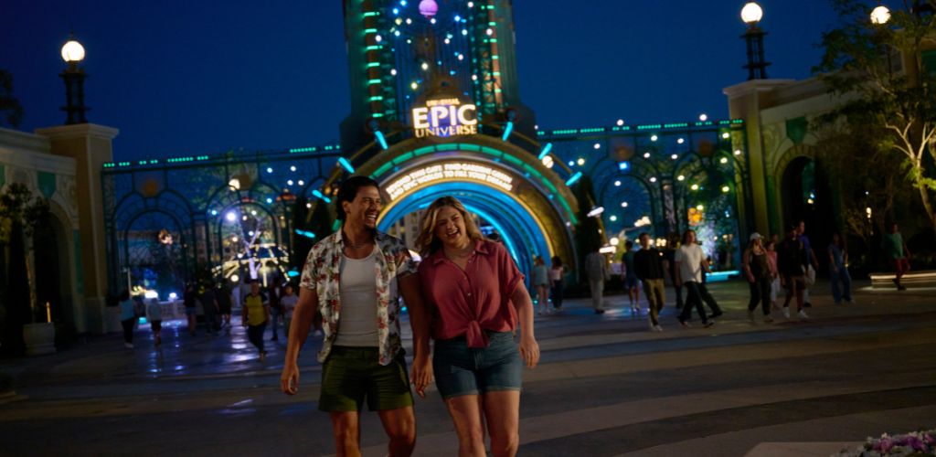 Two people enjoy a nighttime walk in front of an amusement park entrance illuminated with colorful lights and a sign that reads EPIC UNIVERSE.