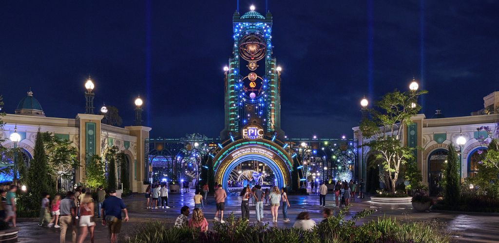 A nighttime view of an illuminated amusement park entrance with colorful lights, a tall central structure, and visitors walking toward the attractions.