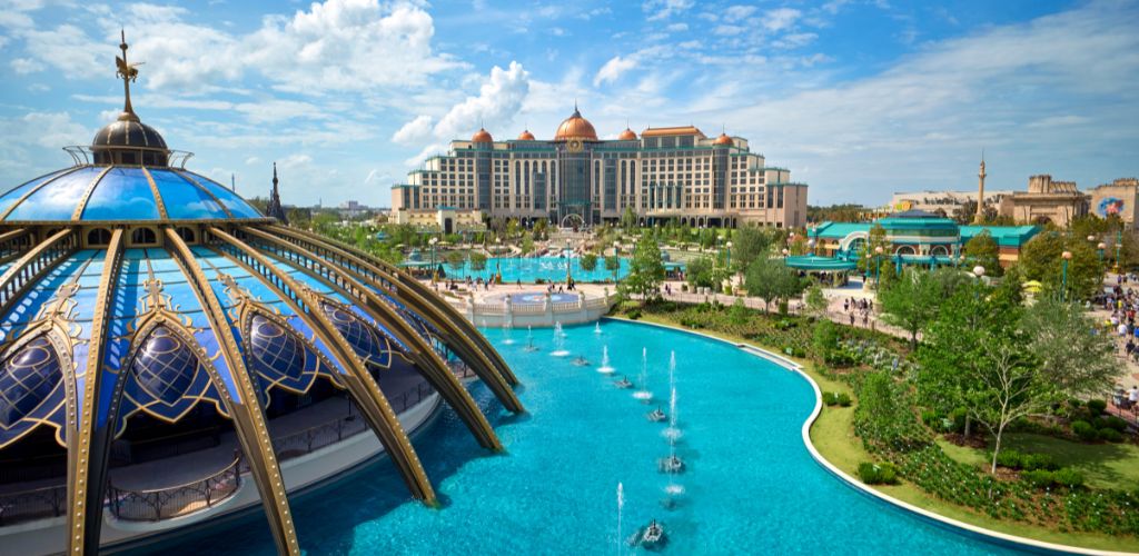 A large theme park with a futuristic building and a domed structure surrounded by water, fountains, and lush greenery under a partly cloudy sky