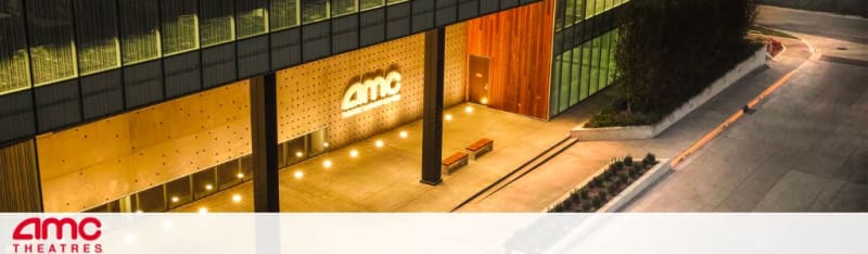 Exterior view at dusk of an AMC Theatres entrance with bright, illuminated signage above the entrance. The facade features warm wood paneling and dotted light fixtures beside a concrete walkway with benches.