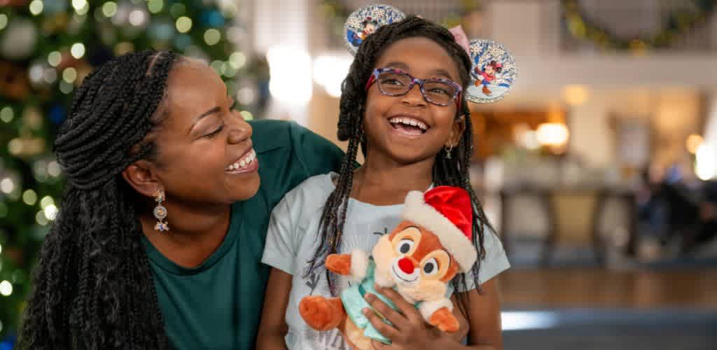 A joyful woman and girl smiling together indoors during the holiday season, girl holds a plush animal wearing a Santa hat.