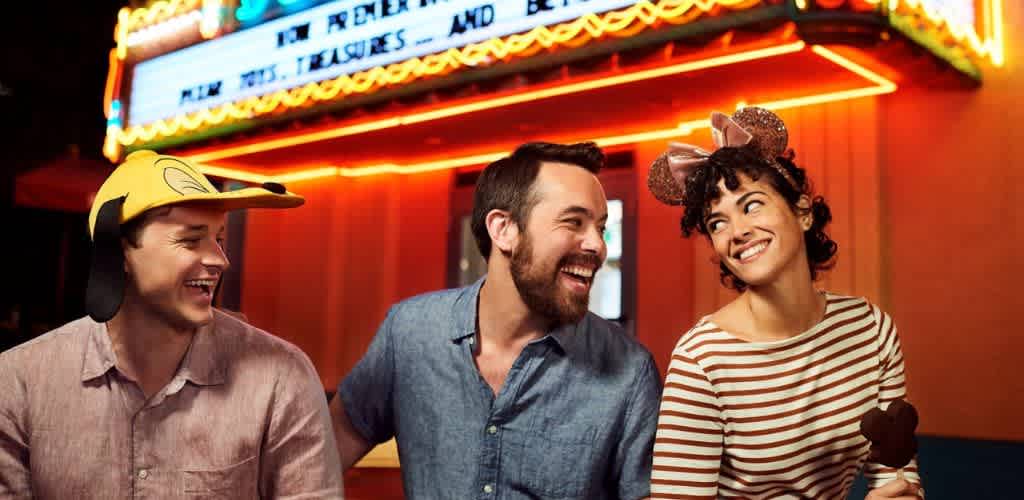 Three people smiling and laughing wearing playful hats in front of a brightly lit marquee at a fun event or party