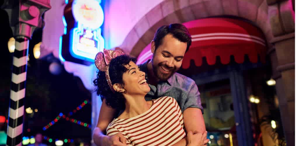 A smiling couple embracing at a colorful, festive indoor party environment with neon lights and holiday decorations.