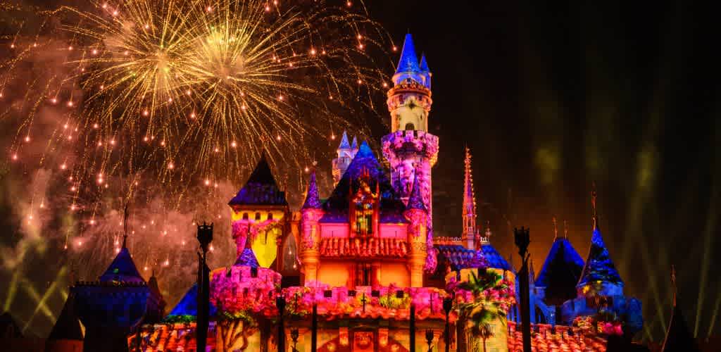 A brightly illuminated fairy tale castle is showcased at night with vibrant projections and colorful lights. In the sky above, golden fireworks burst into brilliant patterns, creating a magical and enchanting atmosphere. The scene exudes a sense of celebration and wonder, perfect for an unforgettable experience for all ages.