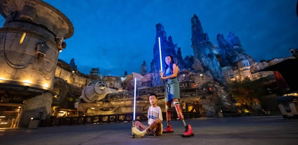 Two people in a Star Wars-themed area hold glowing lightsabers. One person stands confidently, wearing a dress with prosthetic legs, while the other sits on the ground. The scene is set against a backdrop of rocky structures and a spaceship, with evening lighting creating an adventurous atmosphere.
