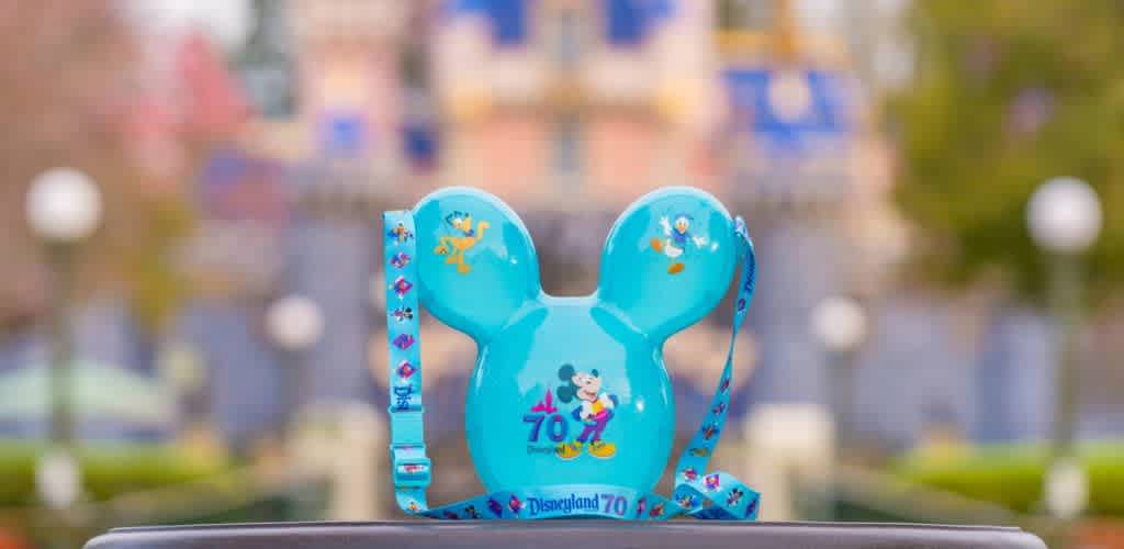 Turquoise Mickey-shaped popcorn bucket celebrates Disneyland's 70th anniversary. It features classic Disney characters and a matching strap with a festive design. The background shows a blurred view of a colorful castle enhancing the whimsical theme. Perfect for fans and collectors.