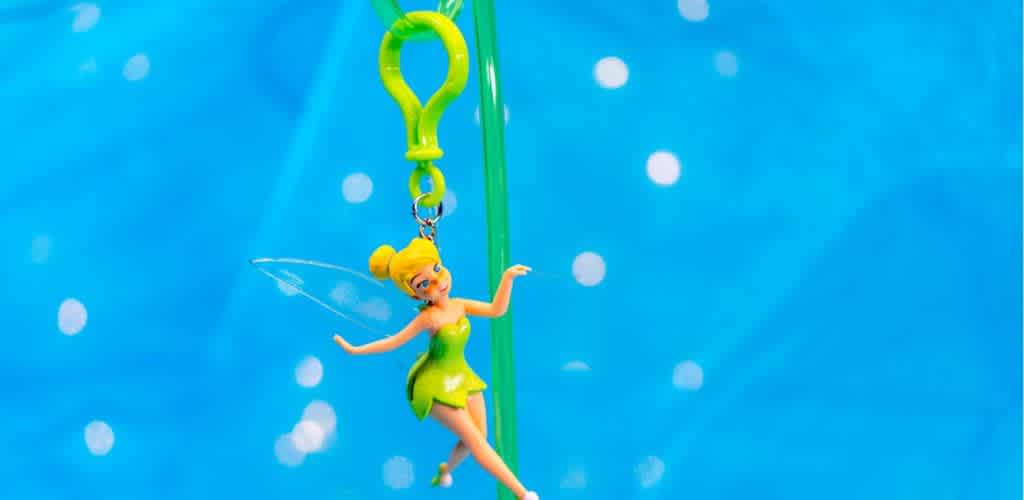 A small figurine of a fairy in a green dress with translucent wings is hanging from a green clip against a blue background with soft white circles. The fairy has blonde hair styled in a bun and is posed gracefully, giving the appearance of flying.