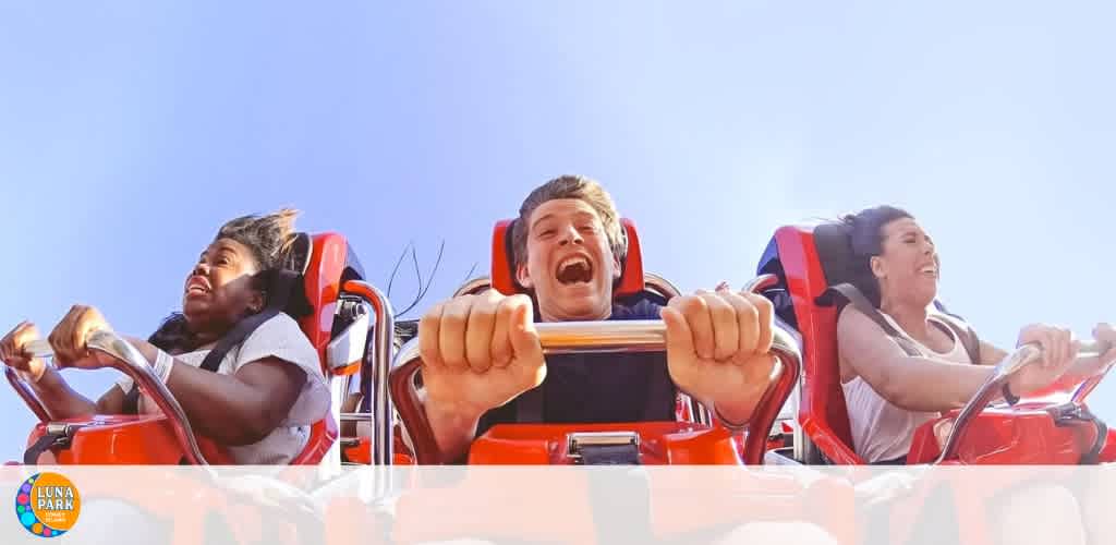 Three people experiencing a thrilling roller coaster ride. They are strapped into a red coaster car, expressing a mix of excitement and fear against a clear blue sky. The first passenger has her mouth open wide as if screaming, the second clenching the safety bar with intense expression, and the third smiling broadly. The Luna Park logo is visible in the corner.