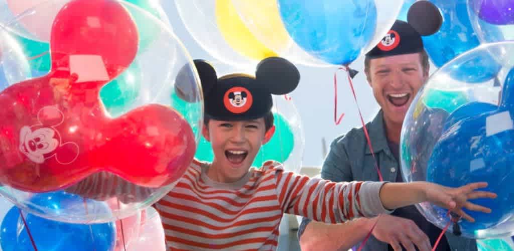 A child and an adult are joyfully holding colorful balloons, both wearing mouse ear hats. The child is in a red and white striped shirt, smiling broadly, while the adult grins beside them. Bright light filters through the balloons, creating a festive and cheerful atmosphere.