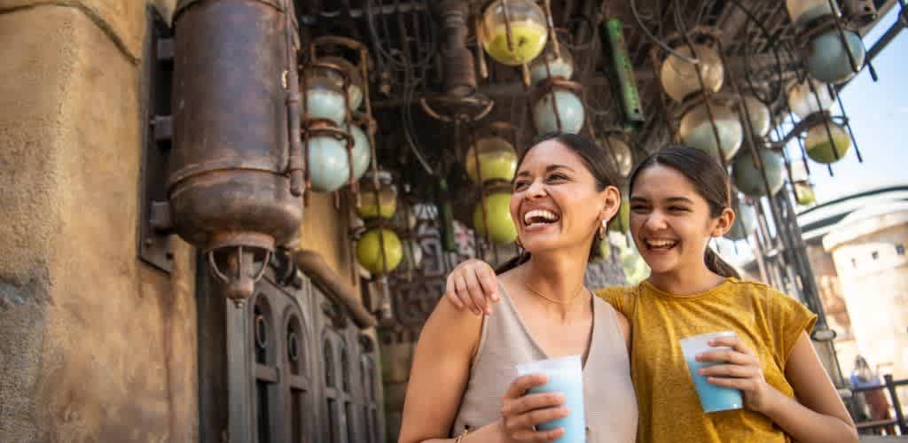 Two people are enjoying a day at an outdoor venue, smiling and holding blue drinks. They stand close together against a backdrop of rustic structures and hanging lights, creating a joyful and vibrant scene. The atmosphere suggests a warm day filled with fun and laughter.