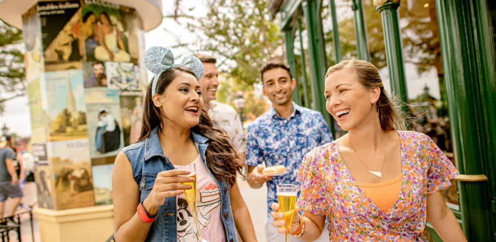 A group of four people joyfully walks outdoors near a green railing and a decorative column adorned with art posters. They are casually dressed, smiling, and chatting, holding drinks in a lively atmosphere. The sky is clear, and trees are visible in the background, suggesting a pleasant day. One person is wearing Mickey Mouse ears, adding a playful touch to the scene.