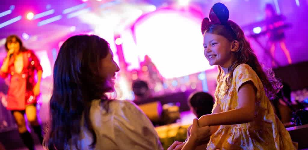A mother and daughter enjoy a lively concert. The mother, wearing a light top, faces her daughter who is smiling and has long hair. The daughter, wearing mouse ears and a yellow dress, looks delighted. A performer in a vibrant pink outfit sings energetically on stage, while colorful lights create an exciting atmosphere.