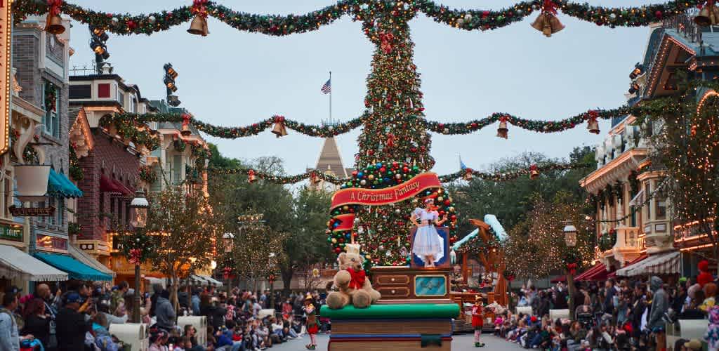 A festive street scene with holiday decorations and a large Christmas tree adorned with ornaments and garlands. A parade float with a person in a blue dress and a large teddy bear passes by. People line the sidewalks, enjoying the festive atmosphere. Colorful storefronts and twinkling lights add to the cheerful setting. The backdrop features a castle with a flag, enhancing the magical ambiance.