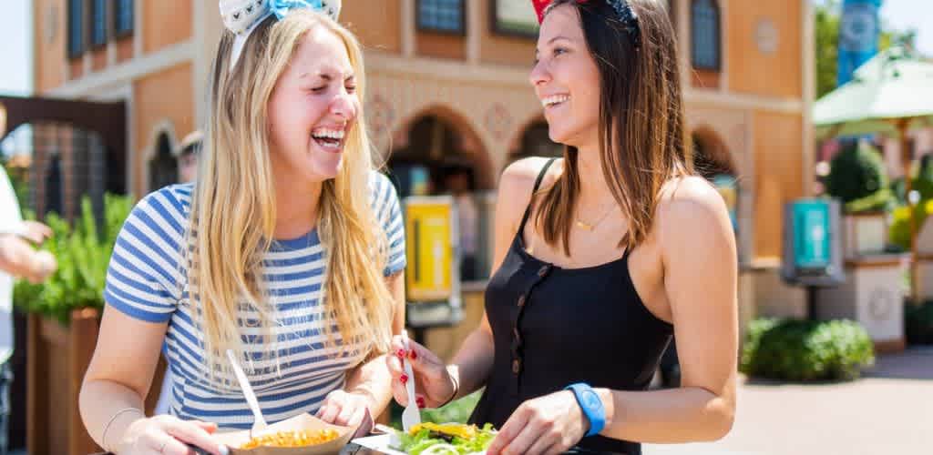 Two women enjoying a meal outdoors at an amusement park. They are laughing and appear to be having a great time. One is holding a bowl of pasta, and the other has a salad. Both wear stylish headbands and casual summer outfits. The background features park buildings, greenery, and a sunny sky, creating a lively atmosphere.