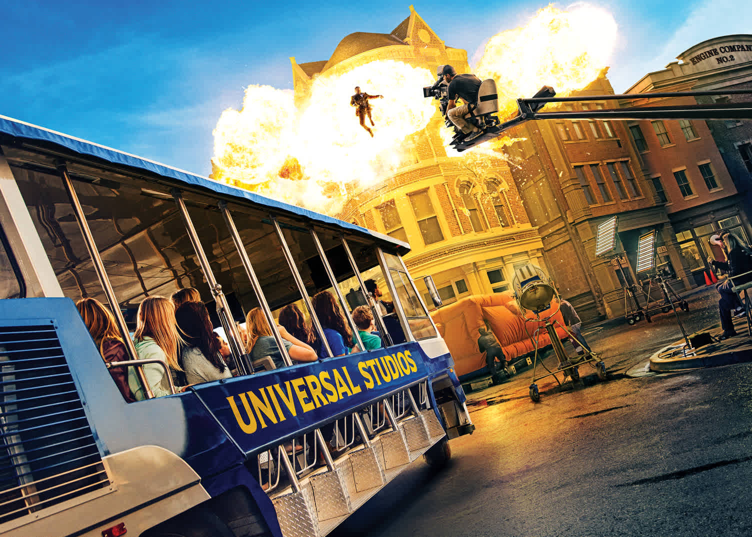 Image of a Universal Studios tour tram filled with passengers looking at a staged action scene. A performer is mid-air amidst fiery explosions against a backdrop of classic building facades. Camera equipment is visible, highlighting the behind-the-scenes movie-making experience.