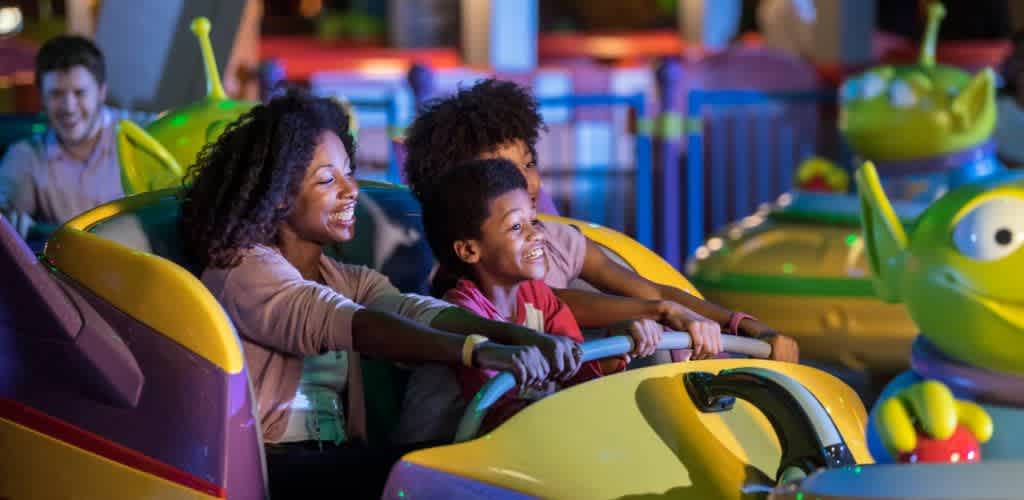 A family enjoys a colorful amusement park ride, smiling and holding onto the safety bar. The scene is lit with vibrant, playful colors, creating an atmosphere of excitement and joy. In the background, other passengers are visible, also enjoying the ride. Mystical creature figures add to the whimsical theme of the attraction.