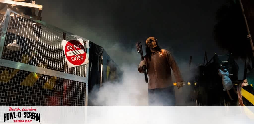 Image shows a promotional scene from Busch Gardens Howl-O-Scream event in Tampa Bay. A figure in a horror costume holds a prop weapon amidst fog. A sign with  Do Not Enter  is visible. Intended for a spooky themed attraction.