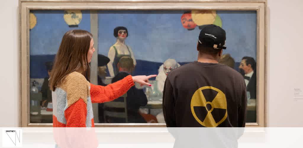 Two visitors are viewing a framed painting in a gallery. The person on the left with long hair is pointing at the painting, wearing a red and grey sweater. The person on the right is facing the artwork, wearing a black cap and grey shirt with a yellow and black circular design. The painting depicts figures seated at a bar.