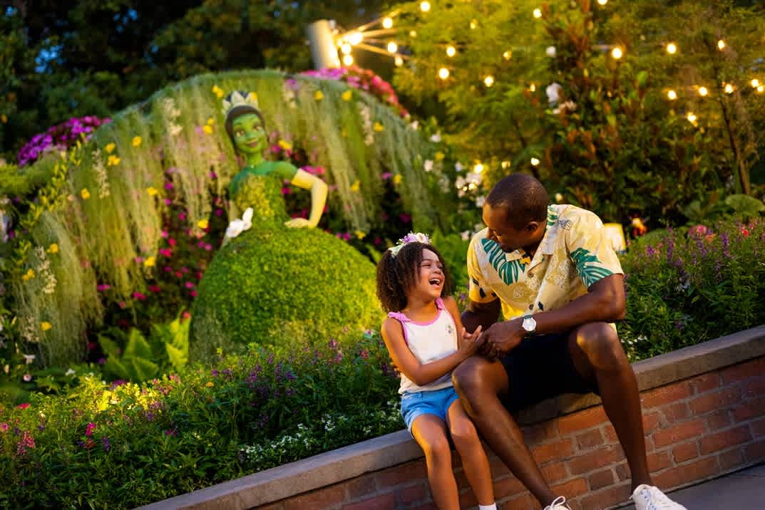 Father and daughter sharing a joyful moment at the EPCOT International Flower & Garden Festival, with a beautifully lit topiary display and vibrant floral scenery in the background.