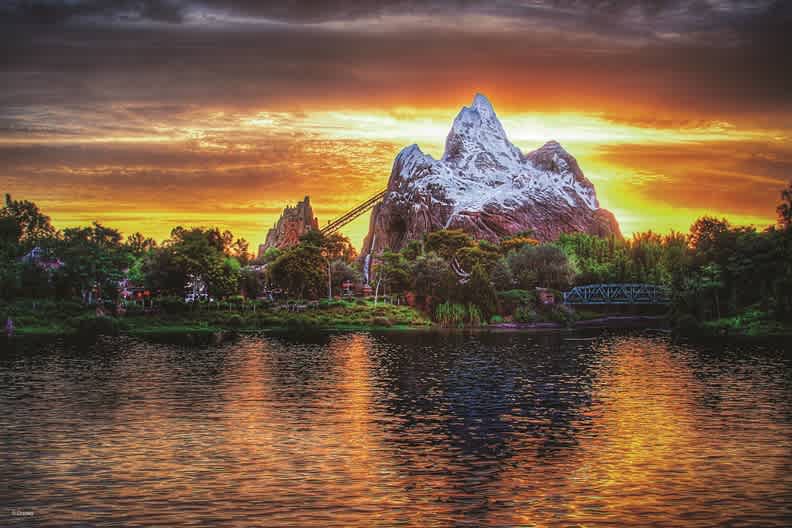 Expedition Everest looms against a breathtaking sunset, its snow-capped peaks reflecting over the water as riders brace for an encounter with the legendary yeti at Disney’s Animal Kingdom.