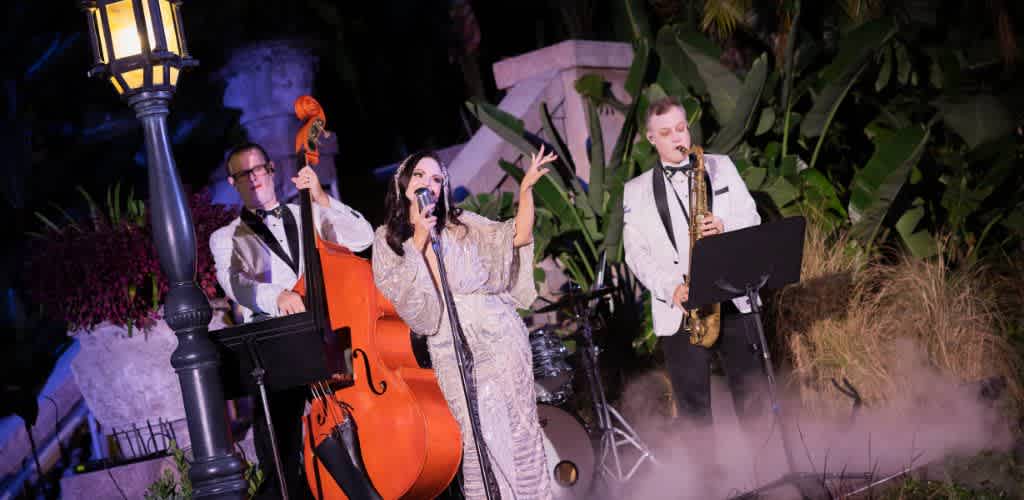 A jazz band performs outdoors at night with lush plants, a street lamp, fog effects, and a woman singing into a microphone.