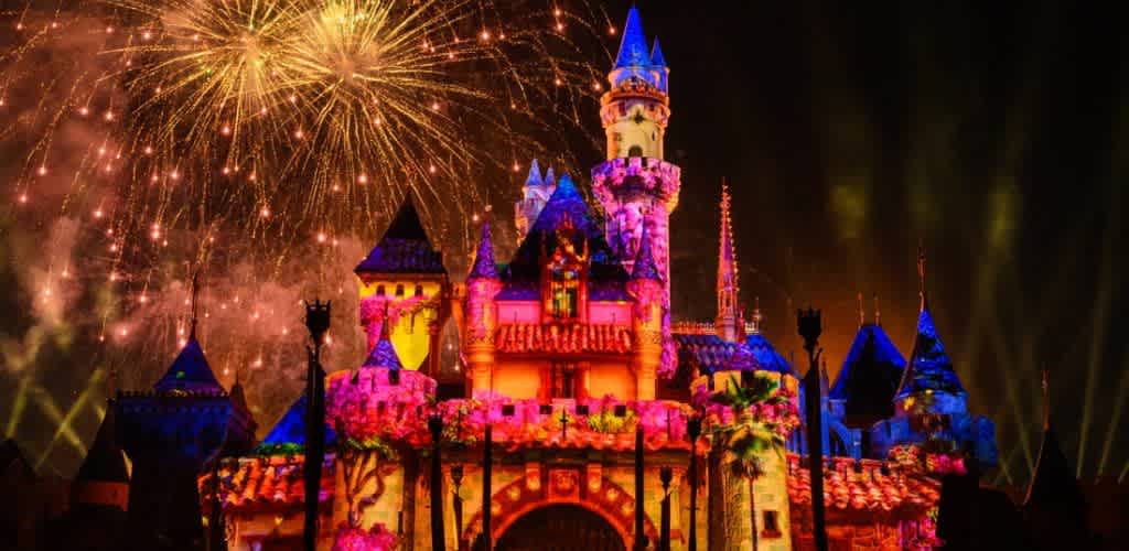 A vibrant nighttime display featuring a brightly lit, colorful castle with fireworks illuminating the sky. The scene captures a magical ambiance with the castle bathed in pink, purple, and blue lights. Fireworks burst above, casting a golden glow and enhancing the enchanting atmosphere. Perfect for a joyful and whimsical experience.