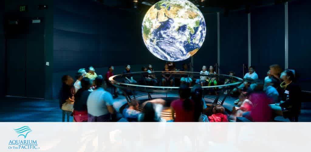 Visitors at the Aquarium of the Pacific gather around a suspended, glowing globe exhibit. They appear engaged with the installation in a dimly lit room accentuating the earth model. The attendees, including children and adults, sit and stand in a semi-circle around the display, some focusing intently on the globe.