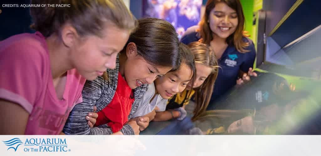 A group of enthusiastic children leans in to observe an exhibit at the Aquarium of the Pacific. They are engaging closely with the display under the guidance of an employee, who is smiling and assisting them. The setting suggests an interactive and educational experience. The image also contains the aquarium's logo and credits the organization for the photo.