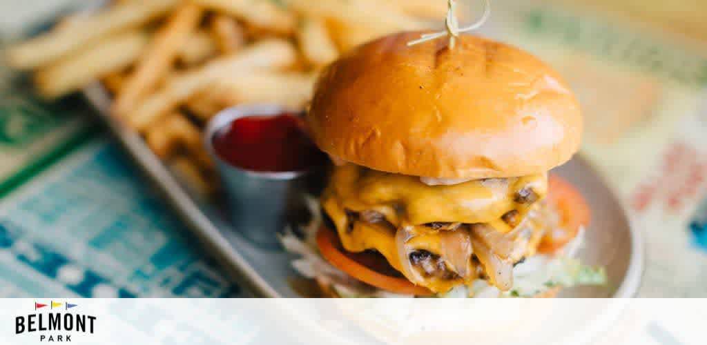 This image shows a close-up of a juicy, double cheeseburger with melted cheese and garnishes, served with a side of fries and ketchup. In the background, a slightly blurred logo reads Belmont Park. The colors are warm and inviting, suggesting a delicious meal.