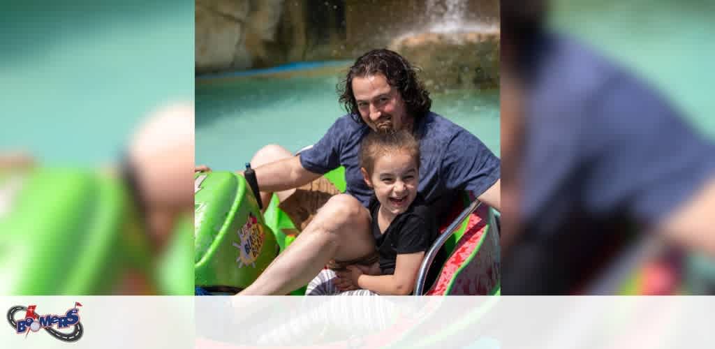 An adult with long hair and a child, both smiling, on a water slide. They are sharing a green inflatable ring. The background shows clear water and a splash, suggesting they are having fun on a sunny day at a water park.