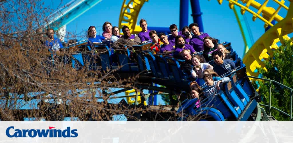 A roller coaster full of excited riders at Carowinds theme park ascends a track with blue and yellow rails against a clear sky. Bare branches in the foreground, thrilling anticipation on faces as they approach the peak.