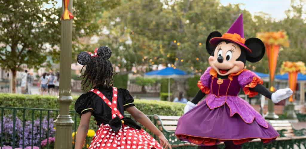 A young girl wearing a Minnie Mouse costume faces a person dressed as Minnie Mouse in a purple Halloween-themed dress outdoors in a park.
