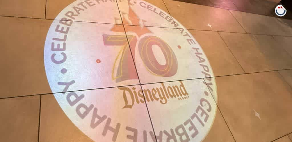 A circular floor projection at Disneyland Resort displays the number 70 in large font, marking a celebration. The surrounding words say Celebrate Happy. The projection sits on a tiled surface, with the area softly lit, creating a festive atmosphere.