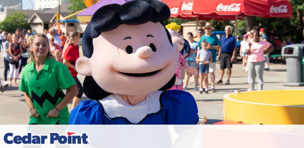 Costumed character in a blue outfit at an amusement park with a crowd and red umbrellas in the background.