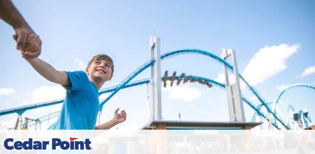 Boy smiling, holding hands with someone, in front of a blue roller coaster.