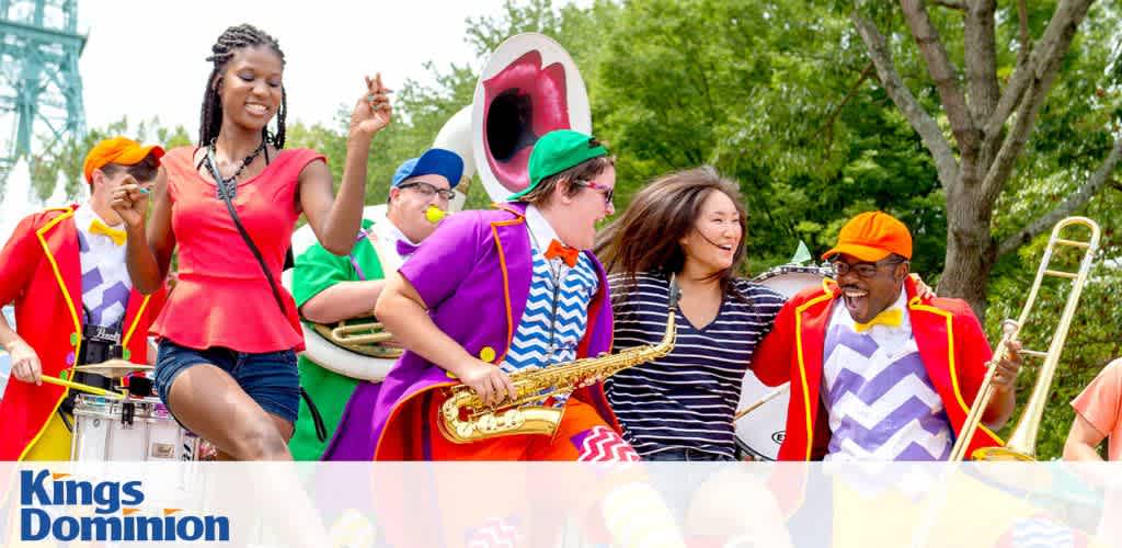 A group of people and clowns in colorful costumes are dancing and playing musical instruments in an outdoor park setting.