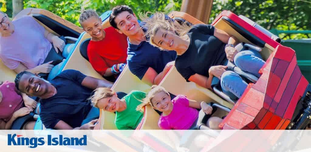 A group of people riding a roller coaster, smiling and laughing with greenery in the background.