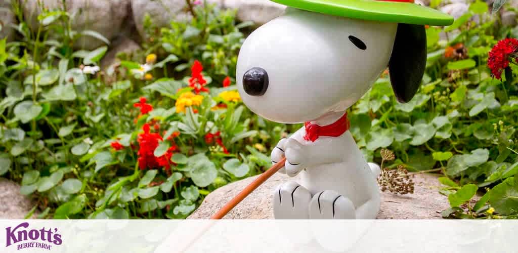 Image shows a statue of the beloved animated character Snoopy as a gardener. He's wearing a green hat, red neckerchief, and holding a watering can, posed as if watering plants. The setting includes red and yellow flowers with greenery in the background. The logo for Knott's Berry Farm is visible in the lower-left corner, indicating the location or theme.