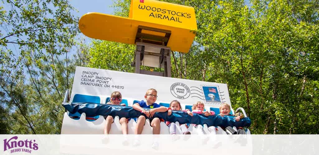 Image shows children enjoying a ride at Knott's Berry Farm. They're seated on a ride that resembles a flying envelope bearing Snoopy's image, poised below a sign saying  Woodstock's Airmail.  It's a sunny day, and trees provide a green backdrop to the fun-filled scene.