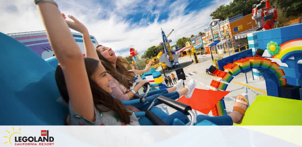 Image of joyful visitors with raised hands on a colorful theme park ride at LEGOLAND California Resort. Brightly hued LEGO-inspired structures and clear skies add to the festive atmosphere.