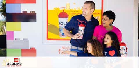 A happy family of four stands in front of a Legoland advertisement showcasing a red cup and a yellow background. The father, wearing a blue shirt, holds the cup while the mother in pink, and two children, appear to be anticipating a fun visit. The Legoland logo is prominent at the bottom.