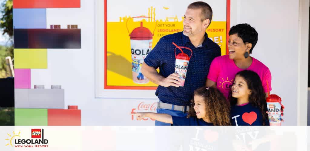 A happy family of four stands before a colorful Legoland-themed backdrop. The father, holding a Legoland cup, looks down smiling at his daughters, while the mother, also smiling, looks at him. The children are joyous, one holding a similar cup. Legoland New York Resort logo is visible.