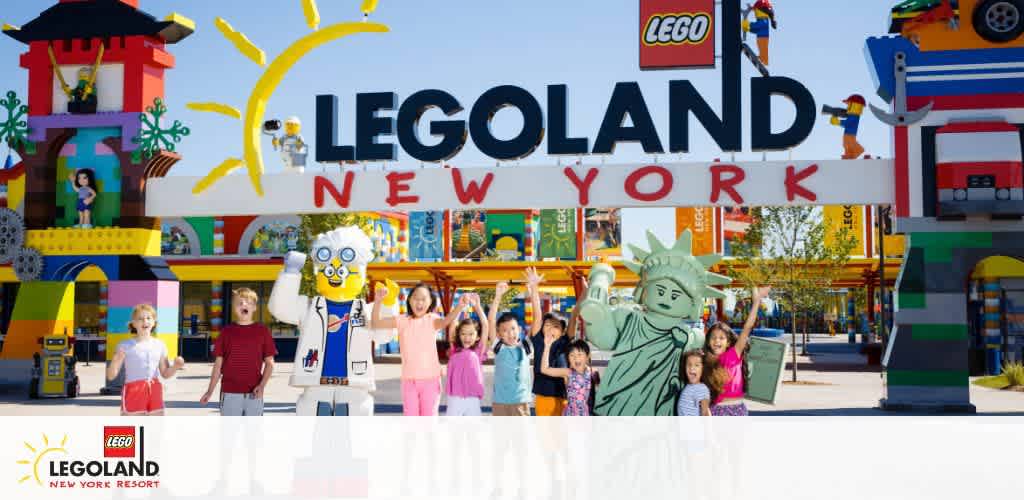 Entrance of LEGOLAND New York Resort with a colorful LEGO-style archway. Excited children are in front, with LEGO figures including a scientist and a Statue of Liberty replica. Clear skies above and LEGO-themed decor surround the joyful scene.